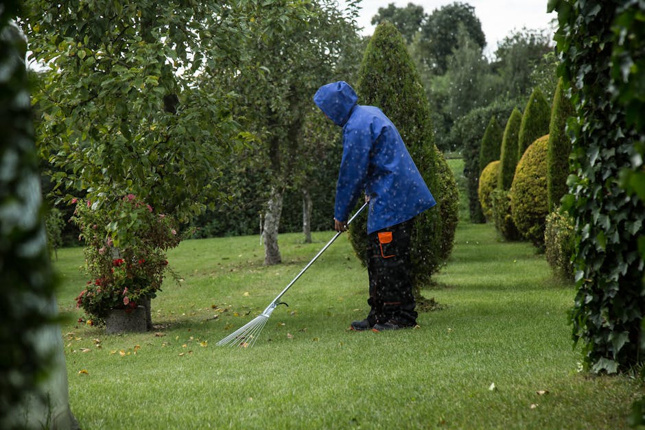 A young woman with light brown hair wearing a beige jacket and white pants is smiling while pushing a lawn mower across a well-maintained grassy garden area. The garden features lush green plants, flo