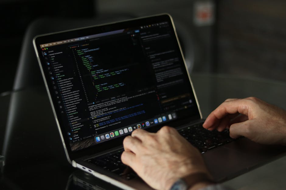 A person typing on a laptop placed on a dark, reflective surface. The laptop screen displays a code editor with lines of colorful programming code on a dark background. The person’s hands are visibl